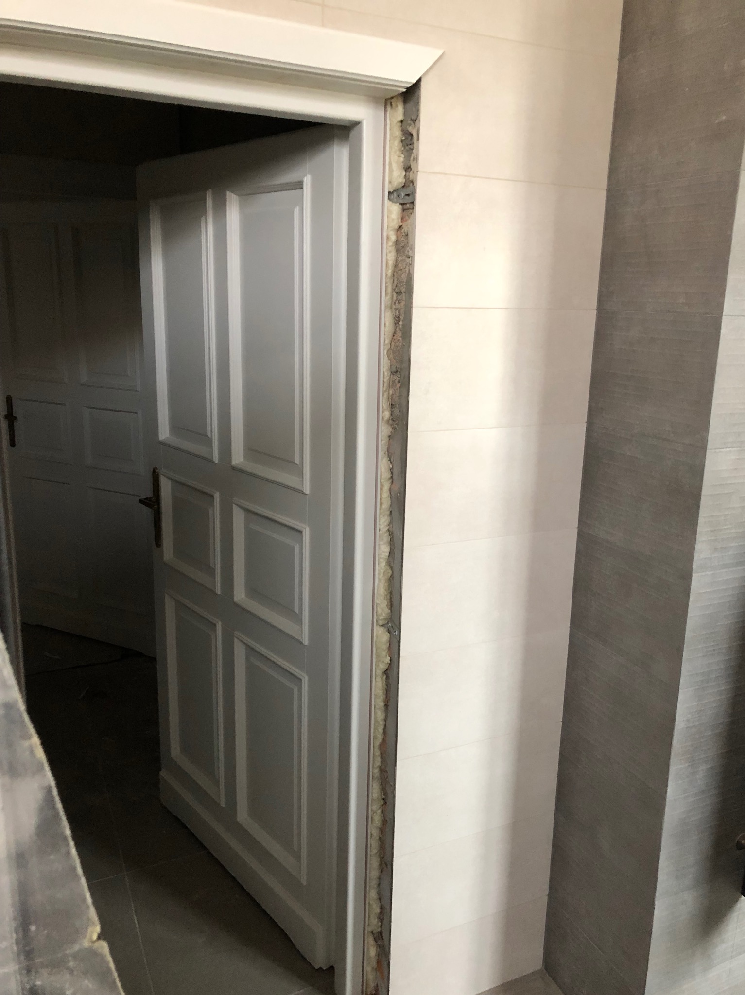 Białe, panelowe drzwi wewnętrzne, częściowo otwarte, z widoczną pianką montażową w ościeżnicy. Tło stanowią białe i szare płytki ścienne.