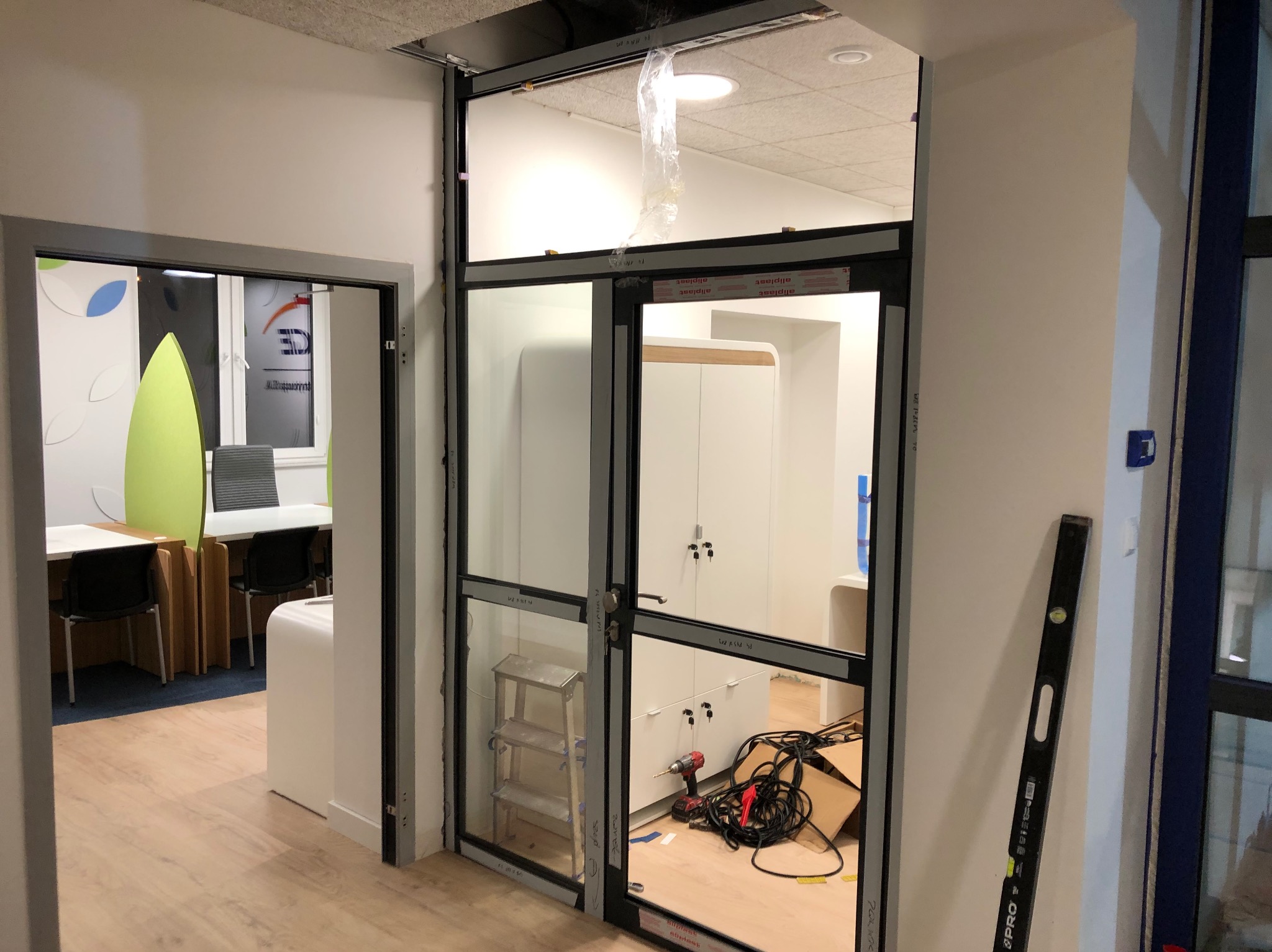 Montaż szklanych drzwi dwuskrzydłowych z czarną ramą. Widoczne narzędzia pracy: poziomica, wiertarka, drabina. W tle biuro z meblami.