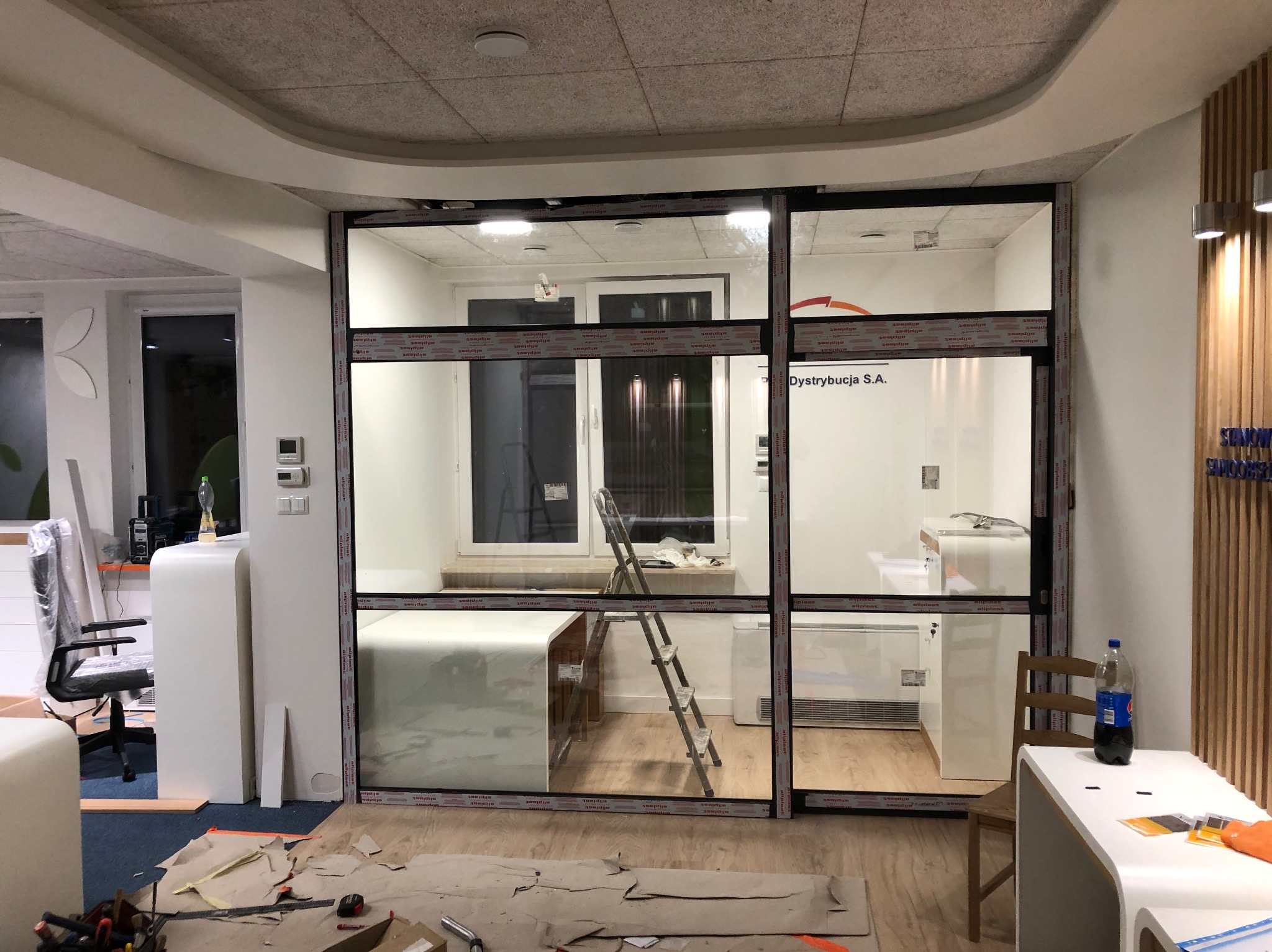 Montaż szklanej ścianki działowej z czarnymi profilami w biurze. Widoczne narzędzia i kartony na podłodze, drabina wewnątrz pomieszczenia. Częściowo widoczne logo producenta profili.