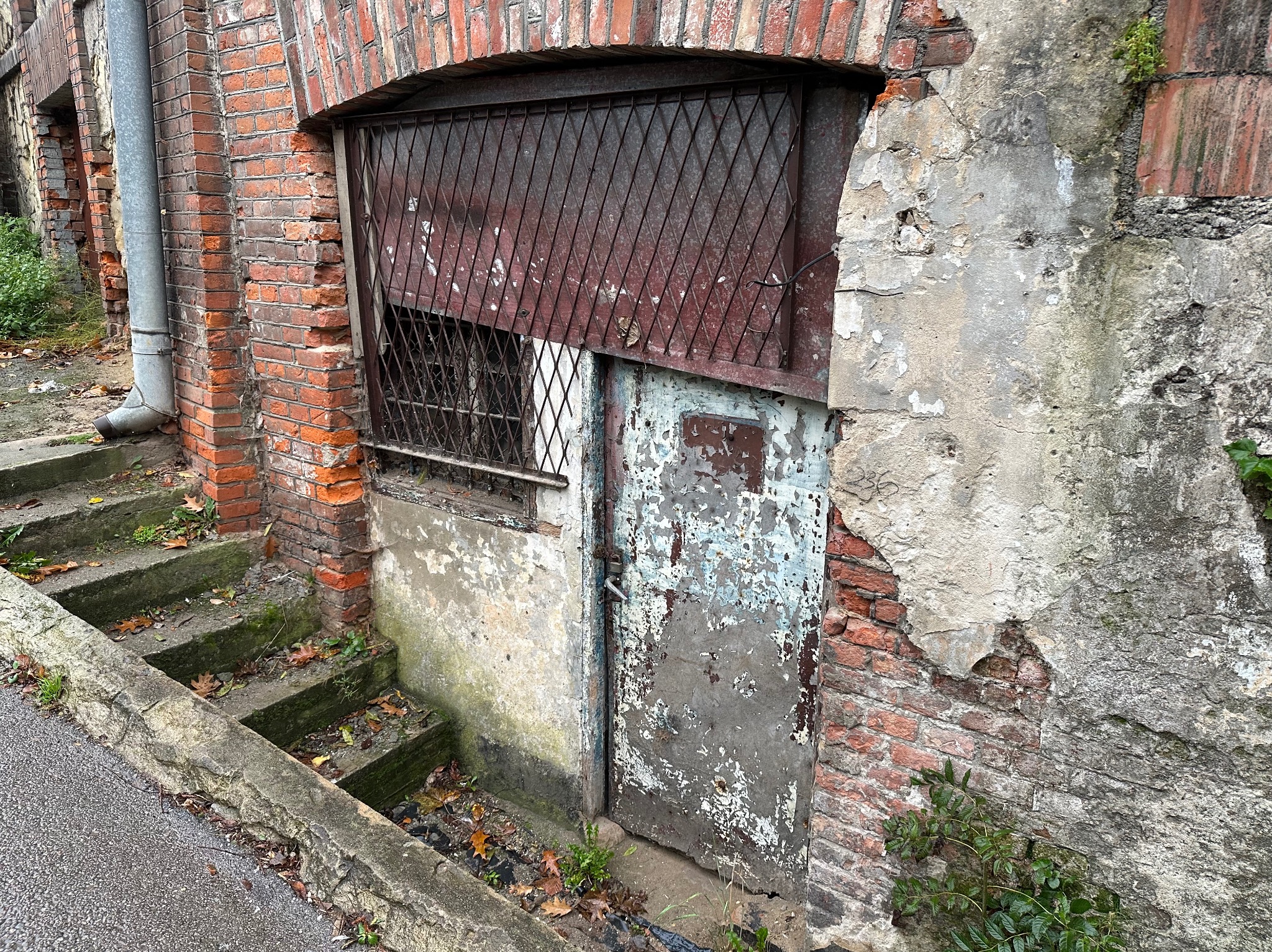 Zrujnowane wejście do piwnicy z metalową kratą i obdrapanymi drzwiami, otoczone ceglanym murem i odpadającym tynkiem. Widoczne schody i rynna.