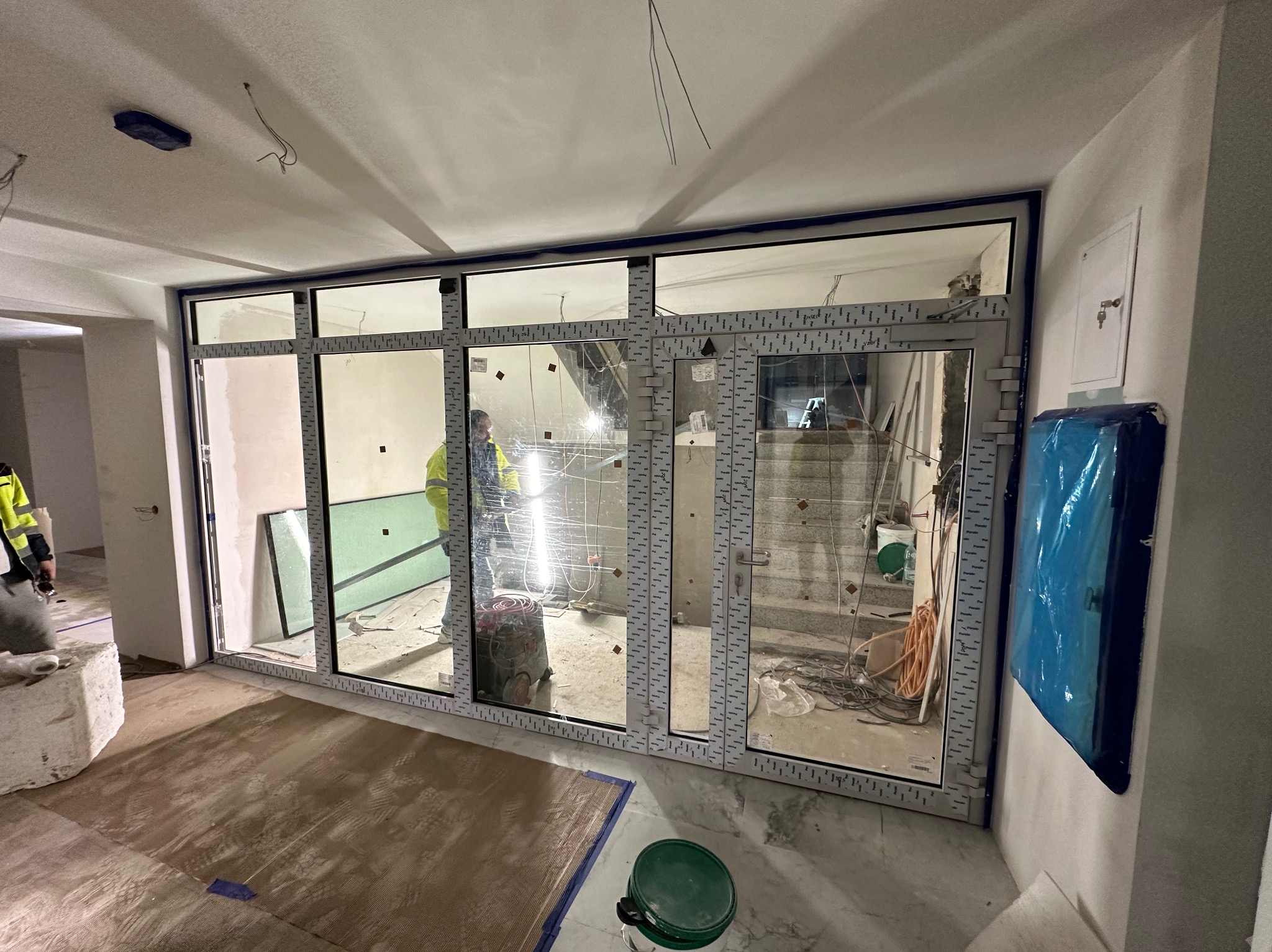 Montaż dużych okien z ramą PCV w surowym wnętrzu. Widoczne narzędzia i elementy wykończeniowe. Praca w toku, z dbałością o detale.