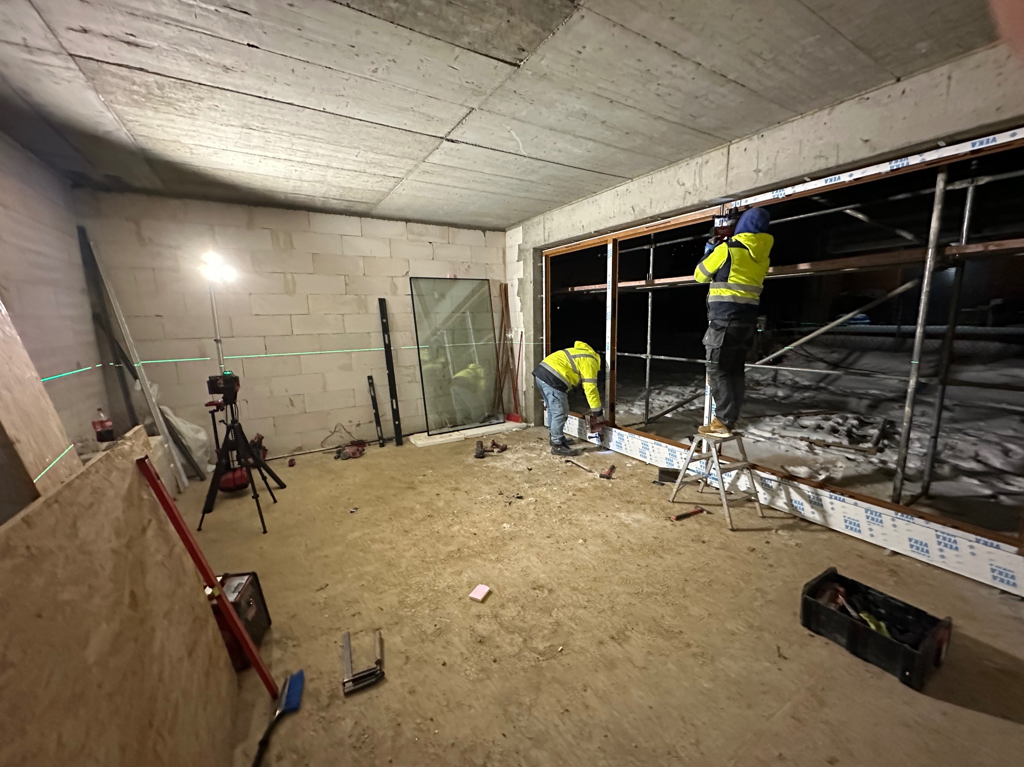 Montaż okna PCV w surowym wnętrzu. Dwóch monterów w żółtych kurtkach pracuje przy oknie, używając poziomicy laserowej. Widoczne narzędzia i materiały budowlane.