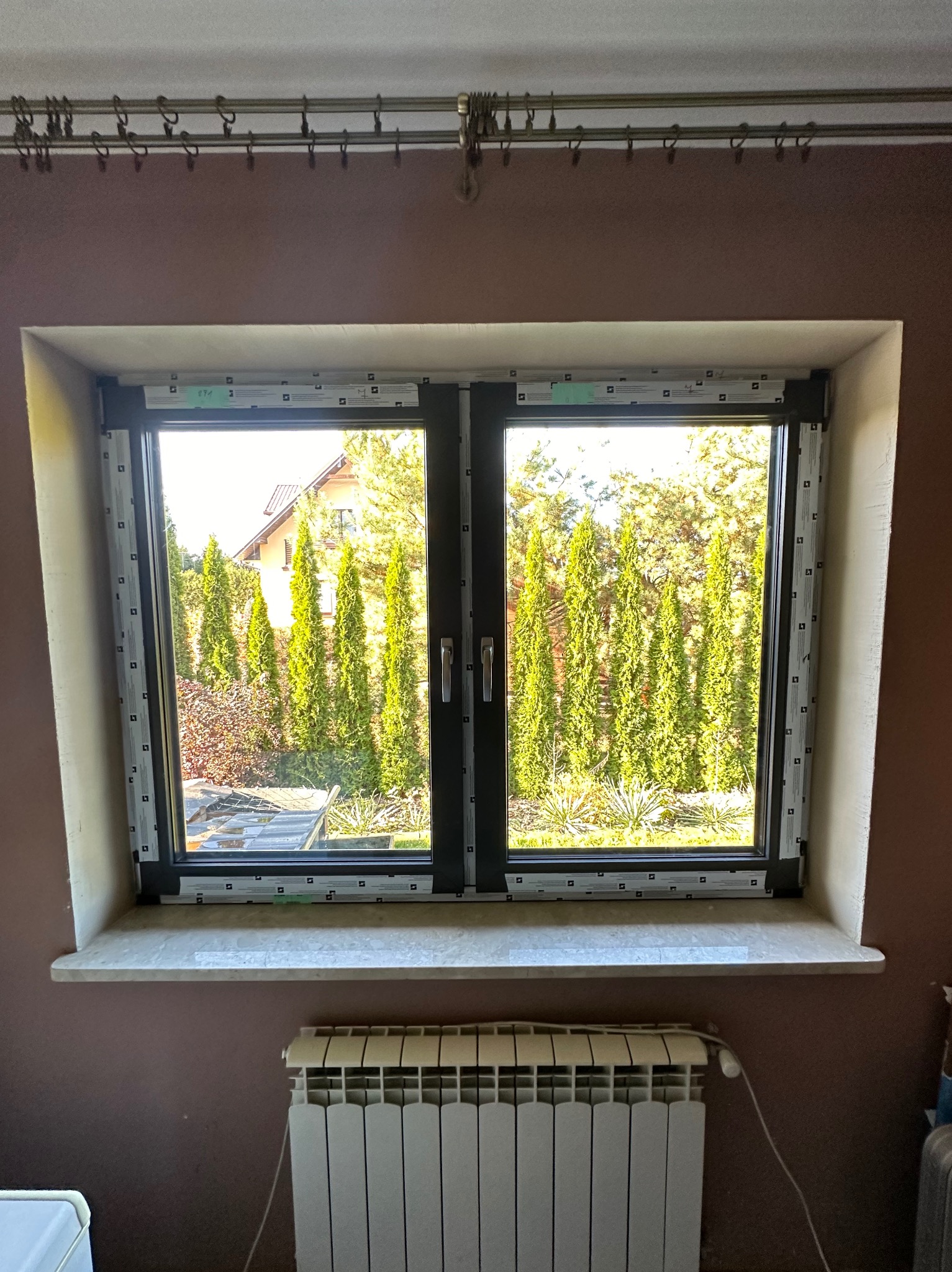 Dwuskrzydłowe okno PCV z czarną ramą, widok na ogród z tujami. Biały parapet i grzejnik pod oknem. Ściana w kolorze brudnego różu.