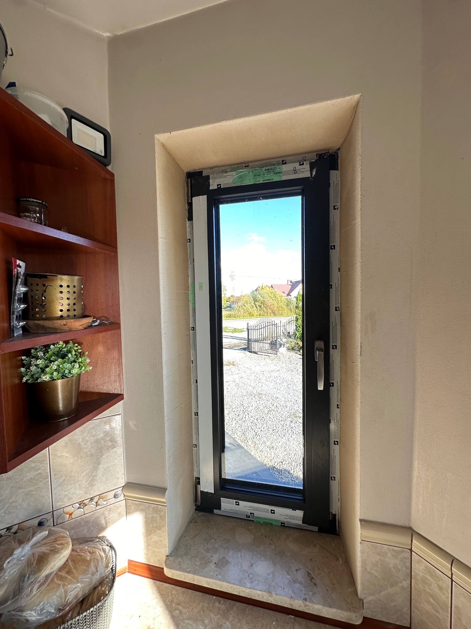 Czarne okno PCV z widokiem na zewnątrz, osadzone w jasnej wnęce. Widoczne detale wykończenia i parapet. W tle półka z dekoracjami i fragment ściany z płytkami.