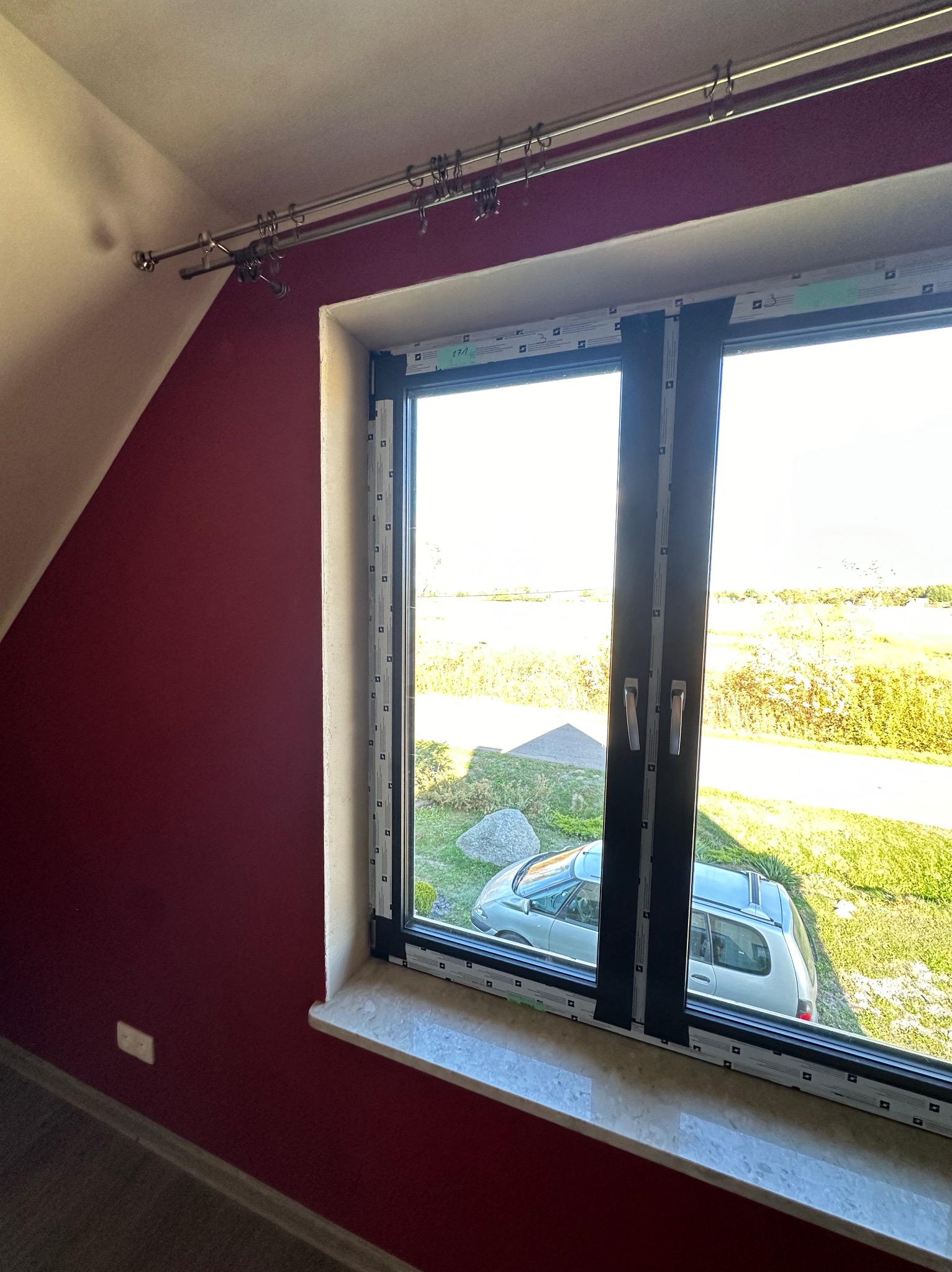 Okno PCV z czarnymi ramami w pokoju na poddaszu z bordową ścianą. Widok na zewnątrz: trawnik, drzewa i zaparkowany samochód. Karnisz na zasłony nad oknem.