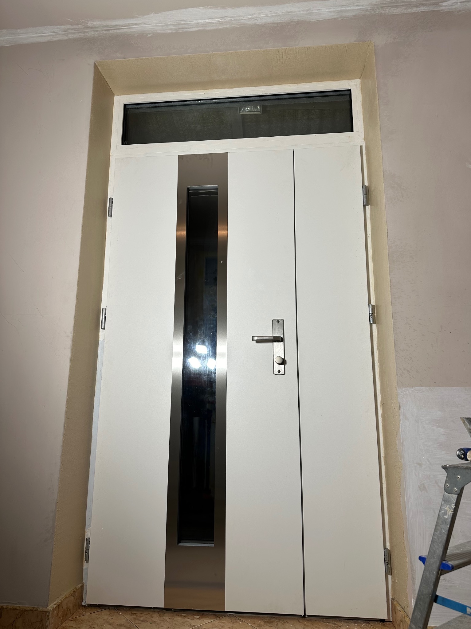 Białe, nowoczesne drzwi wewnętrzne z przeszkleniem i metalowymi akcentami, zamontowane w ościeżnicy. Widoczne ślady prac wykończeniowych na ścianach i drabina.