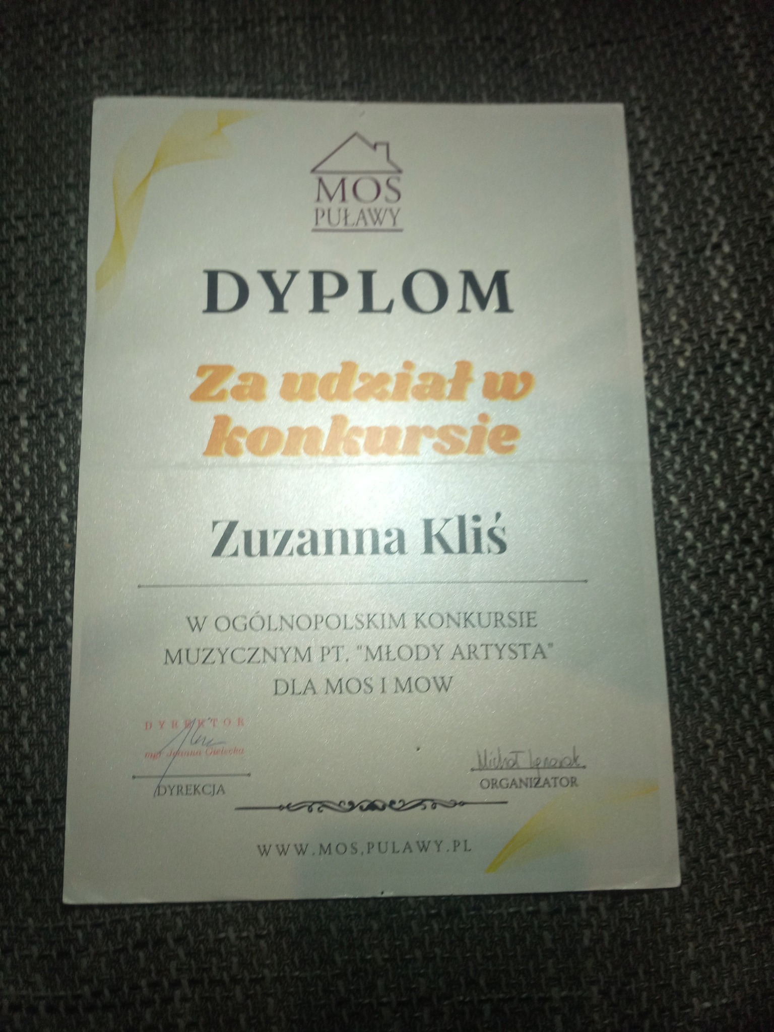 Dyplom za udział w konkursie muzycznym 'Młody Artysta' dla Zuzanny Kliś, wydany przez MOS Puławy. Dokument leży na szarej powierzchni.