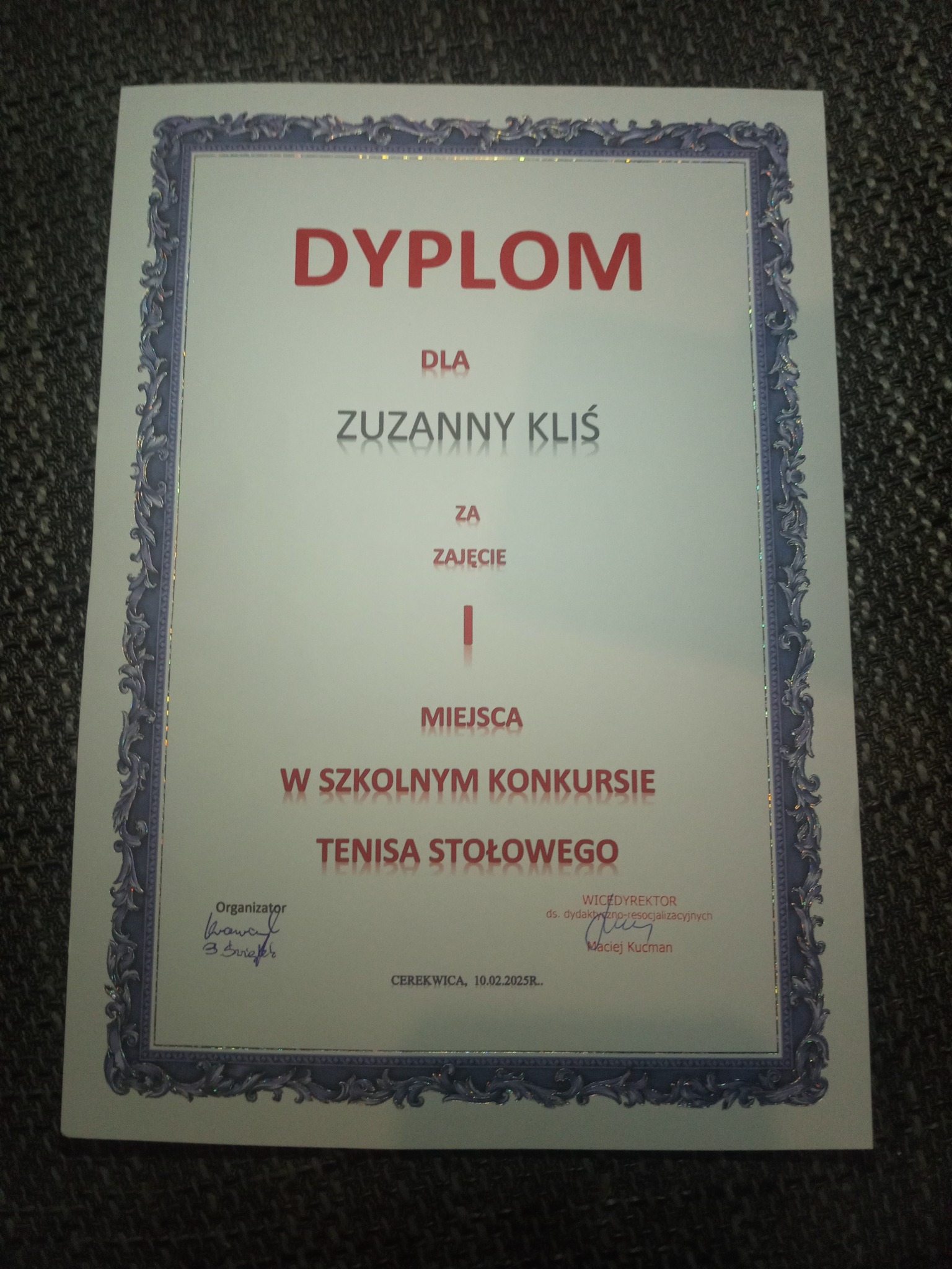 Dyplom dla Zuzanny Kliś za zajęcie miejsca w szkolnym konkursie tenisa stołowego, z ozdobną ramką i podpisami organizatorów. Dokument leży na szarym tle.