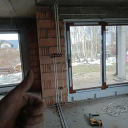 Instalacja elektryczna w surowym wnętrzu: rury prowadzone po ścianie z cegły, puszki elektryczne. W tle okno z widokiem na zimowy krajobraz. Kciuk w górę.