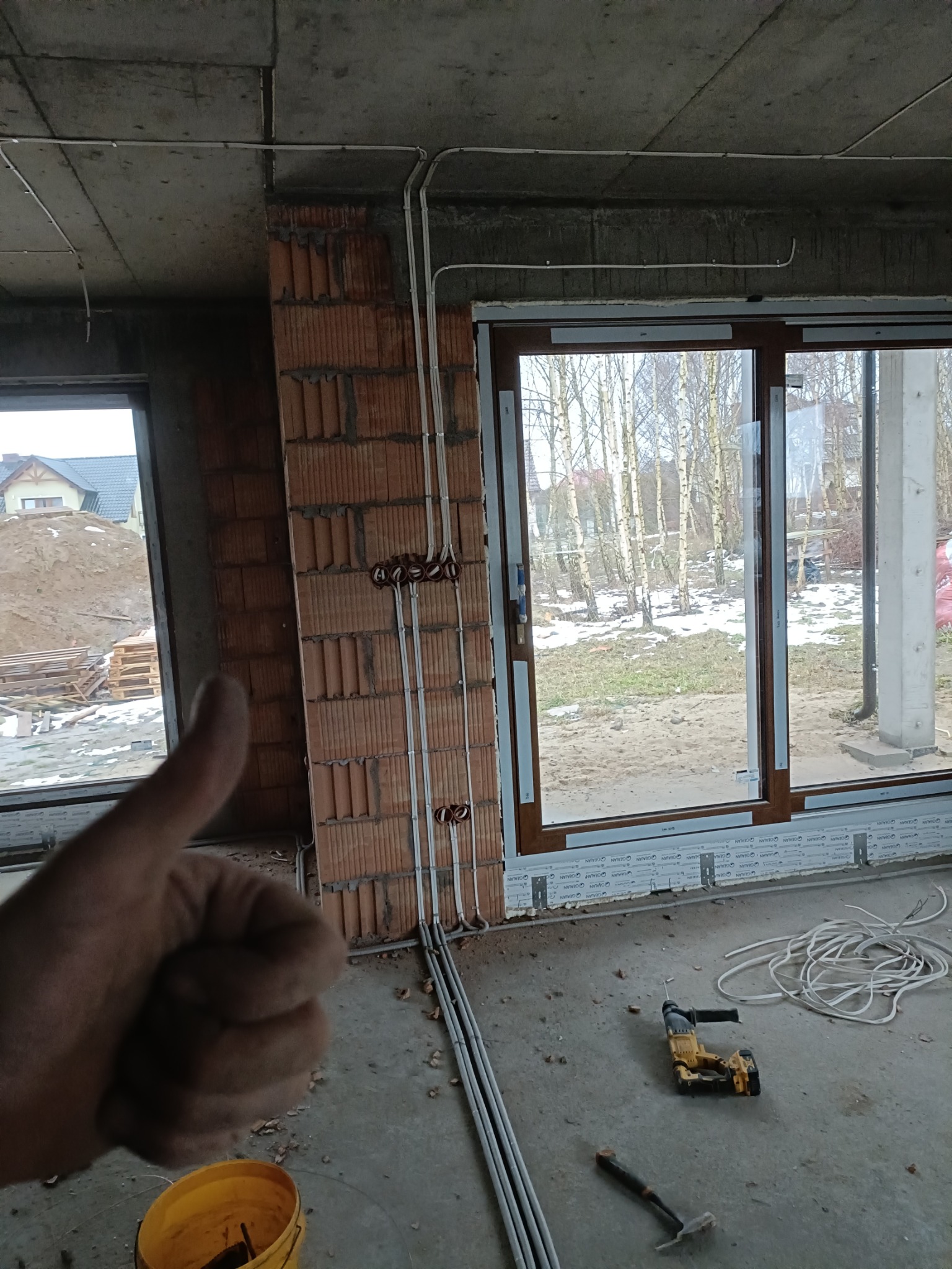 Instalacja elektryczna w surowym wnętrzu: rury prowadzone po ścianie z cegły, puszki elektryczne. W tle okno z widokiem na zimowy krajobraz. Kciuk w górę.