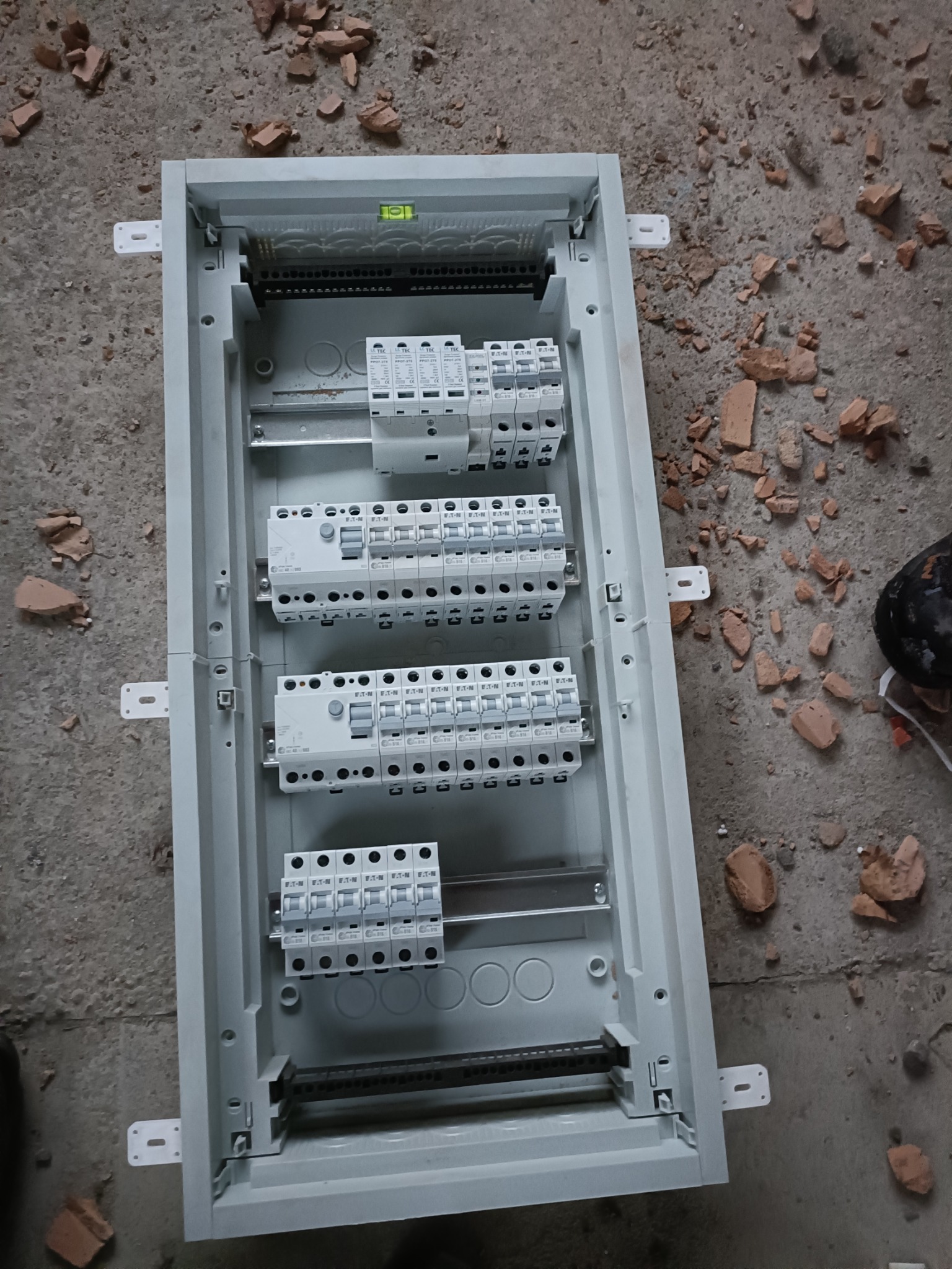 Rozdzielnia elektryczna w trakcie montażu, widoczne zabezpieczenia i listwy, poziomica na górze. Otoczenie budowlane, gruz na podłodze.