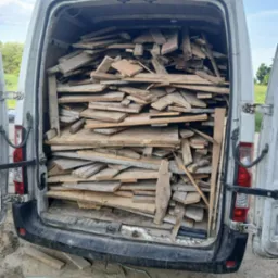 Otwarty bagażnik białego busa wypełniony po brzegi różnymi kawałkami drewna, desek i płyt. Widoczne otoczenie wiejskie: pole i drzewa.