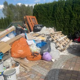 Mateusz Majkowski - Stos odpadów budowlanych i poremontowych na kostce brukowej, worki, gruz, paleta, drzwi, w tle zielony żywopłot i drzewa. Słoneczny dzień.