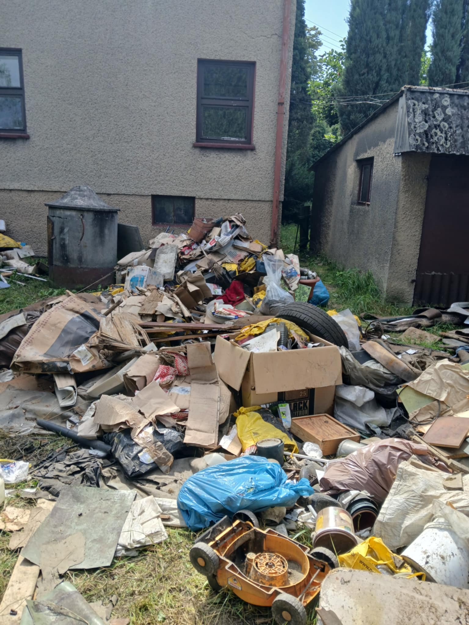 Stary, zaniedbany ogród z nagromadzeniem śmieci, w tym kartony, opony i zabawki, z budynkiem w tle. Widoczny zaniedbany trawnik i elementy wyposażenia ogrodu.