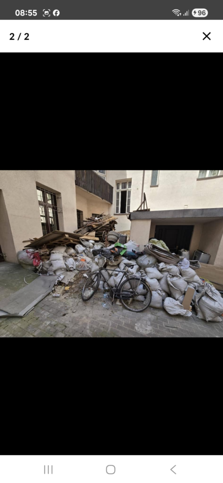 Stare podwórko kamienicy zawalone workami gruzu, stertą desek i starym rowerem opartym o stertę śmieci. Widoczne okna i balkon budynku w tle.