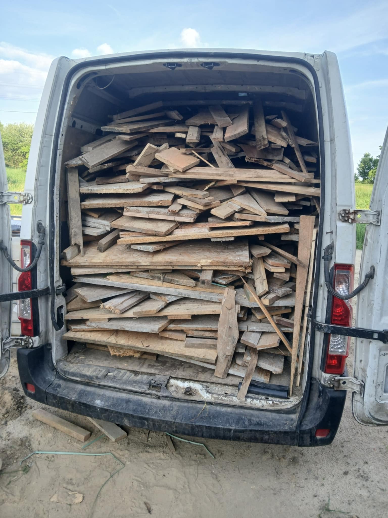 Otwarty bagażnik białego busa wypełniony po brzegi różnymi kawałkami drewna, desek i płyt. Widoczne otoczenie wiejskie: pole i drzewa.