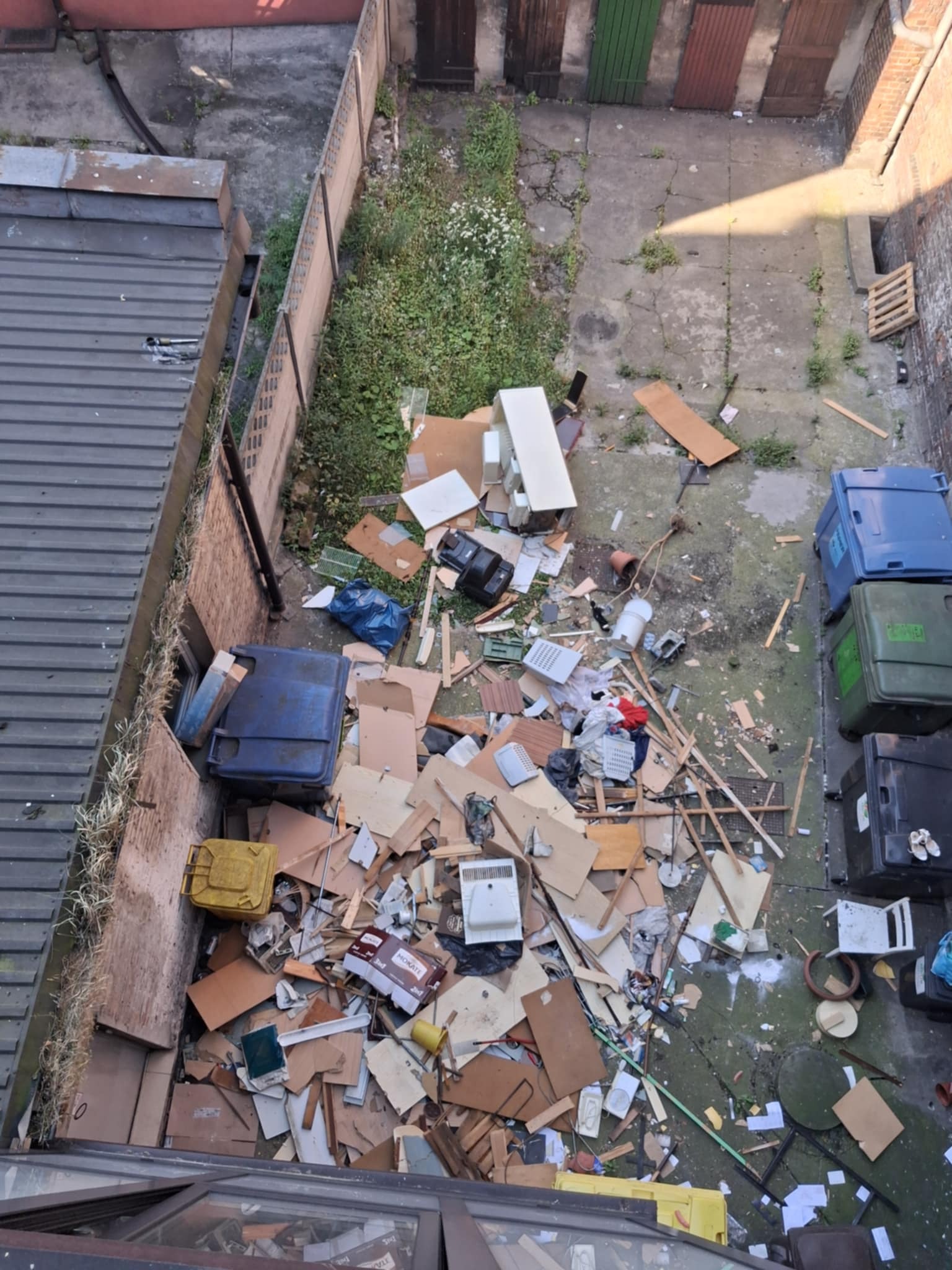 Widok z góry na podwórko z gruzem i odpadami, obok stoją kontenery na śmieci. Widoczne są stare meble i inne przedmioty. Częściowo zarośnięty teren.