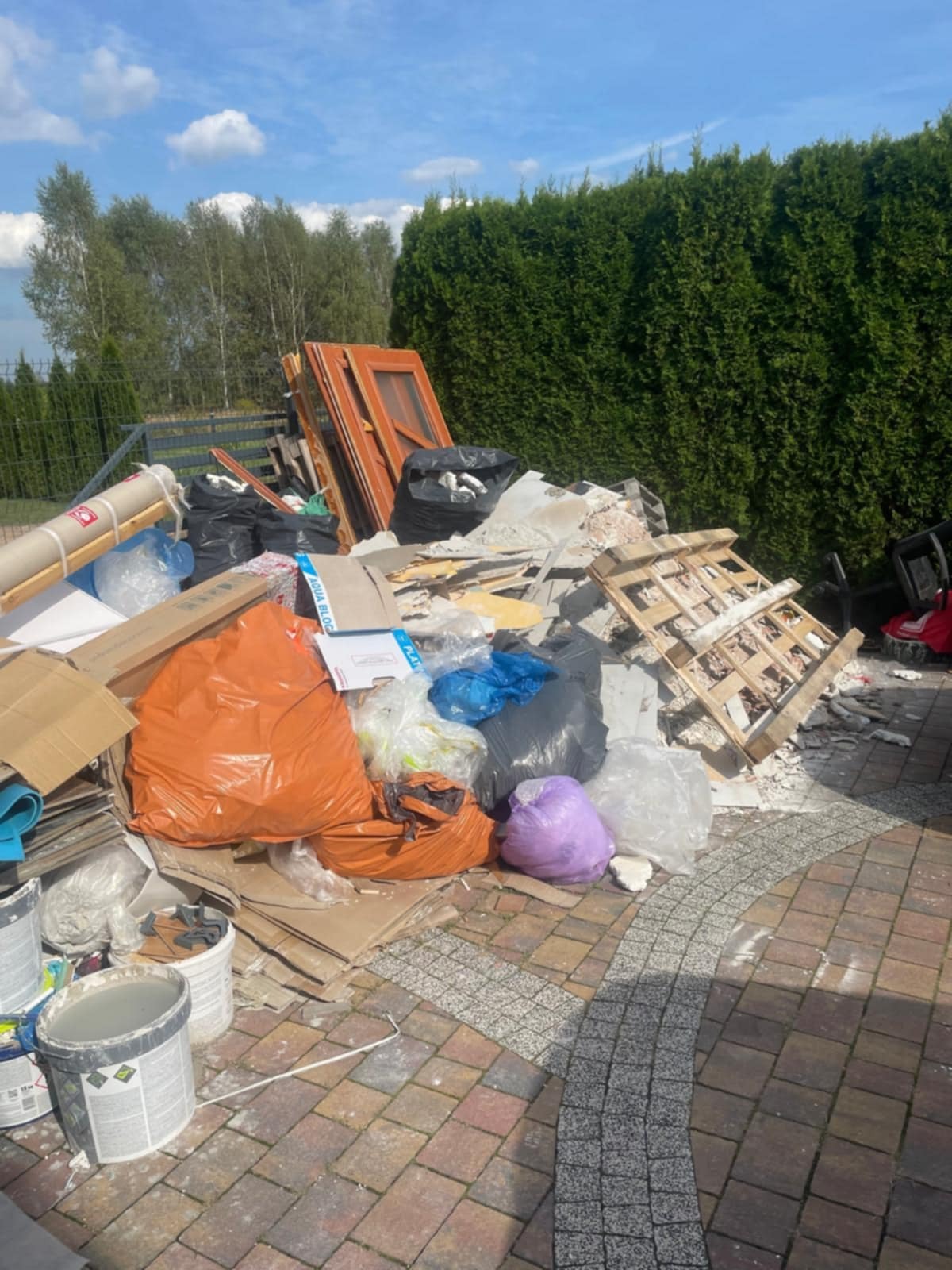 Stos odpadów budowlanych i poremontowych na kostce brukowej, worki, gruz, paleta, drzwi, w tle zielony żywopłot i drzewa. Słoneczny dzień.