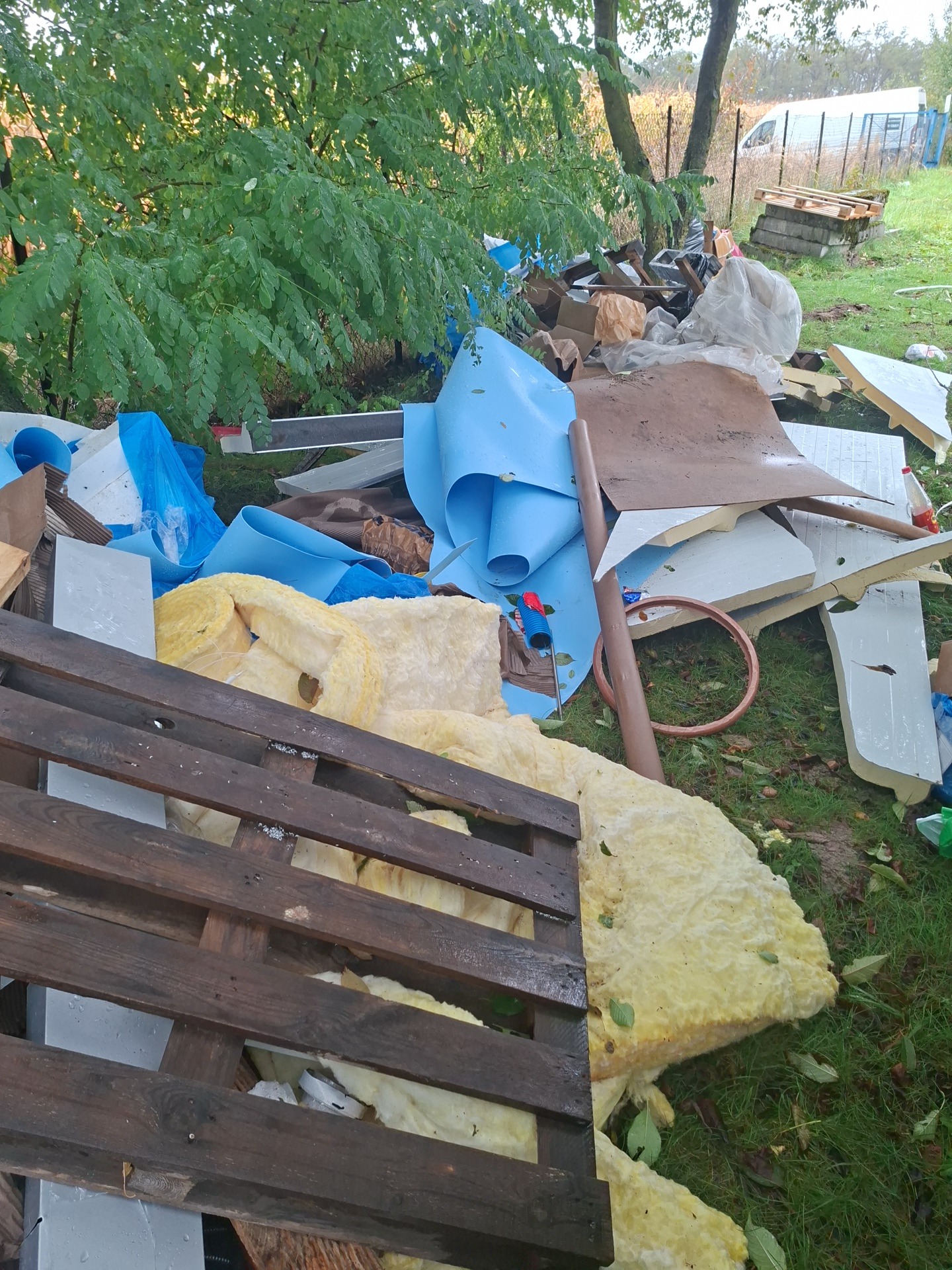 Sterta odpadów budowlanych i gruzu na trawie, w tle zieleń i częściowo widoczny biały bus. Widoczne elementy izolacji, paleta i fragmenty płyt.