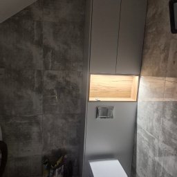 Ytsenko Dmytro - Szara szafka wnękowa z drewnianą półką podświetloną LED nad toaletą w łazience wykończonej płytkami imitującymi beton, widoczne narzędzia glazurnicze.