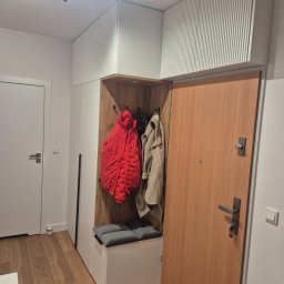 Ytsenko Dmytro - Nowoczesna zabudowa przedpokoju z szafą z ryflowanym frontem, siedziskiem i wnęką na wieszaki. Widoczne ubrania i drewniane wykończenie wnęki. Jasne wnętrze.