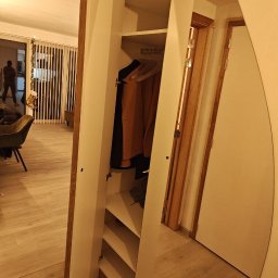 Ytsenko Dmytro - Biała szafa wnękowa z lustrem, częściowo otwarta, ukazująca półki i wiszące ubrania. Widoczne drewniane wykończenia i jasne wnętrze. Perspektywa z wnętrza mieszkania.