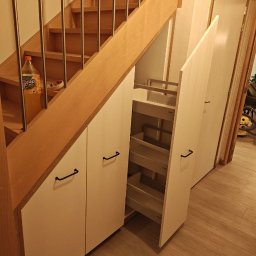 Ytsenko Dmytro - Biała zabudowa pod schodami z wysuwanymi półkami, czarne uchwyty. Schody drewniane z metalową balustradą. Na schodach napój w plastikowej butelce.