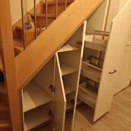 Ytsenko Dmytro - Szafka wnękowa pod schodami z białymi frontami, otwartymi drzwiczkami i wysuwanymi półkami. Na schodach stoi butelka napoju.