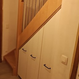 Ytsenko Dmytro - Biała szafa wnękowa pod schodami z drewnianą balustradą i czarnymi uchwytami. Widoczny fragment schodów i włącznik światła na szafie.