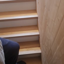 Ytsenko Dmytro - Drewniane schody z białymi podstopnicami, widok z dołu, fragment osoby w kadrze. Naturalne oświetlenie podkreśla strukturę drewna i detale wykończenia.