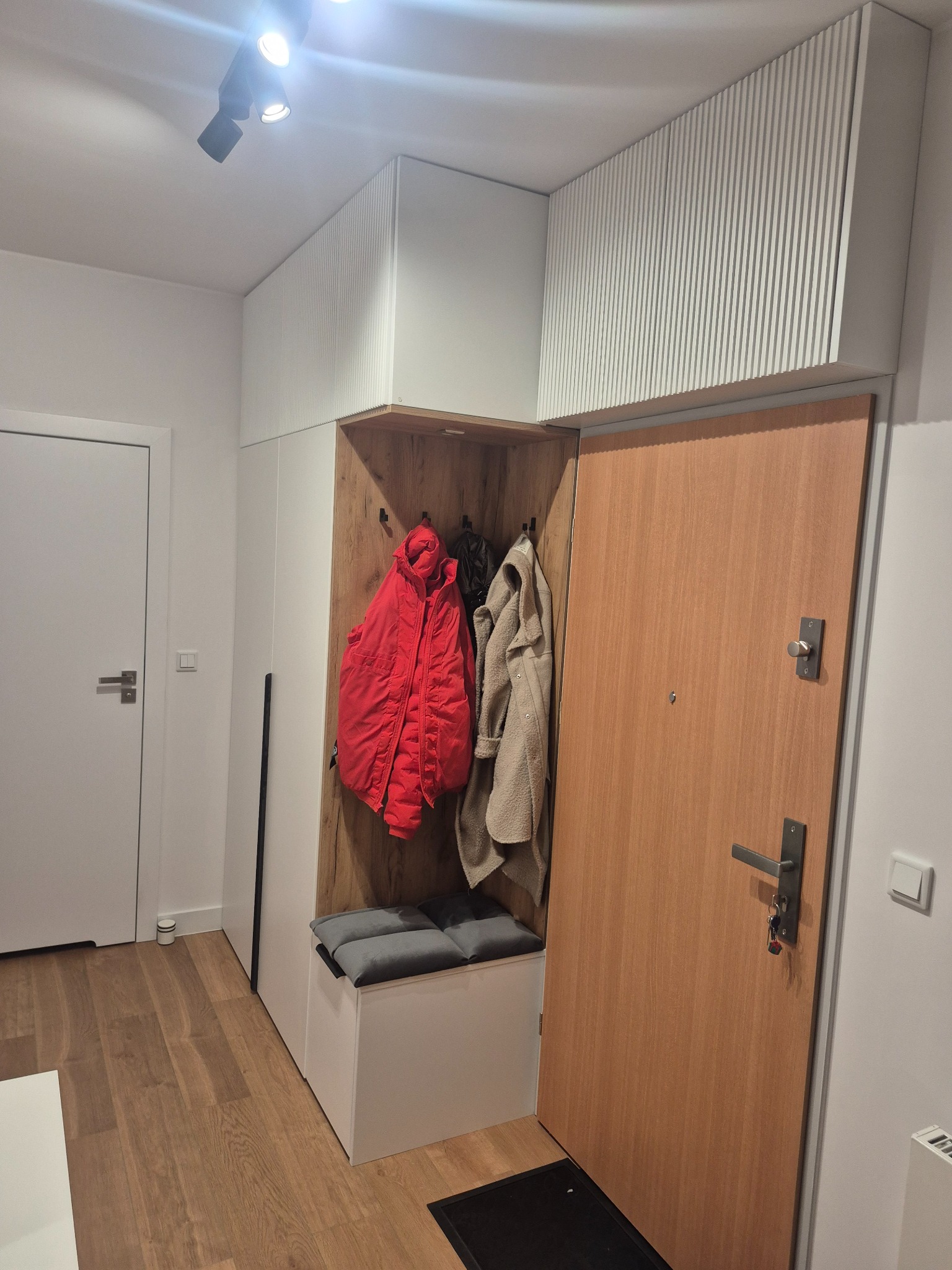 Nowoczesna zabudowa przedpokoju z szafą z ryflowanym frontem, siedziskiem i wnęką na wieszaki. Widoczne ubrania i drewniane wykończenie wnęki. Jasne wnętrze.