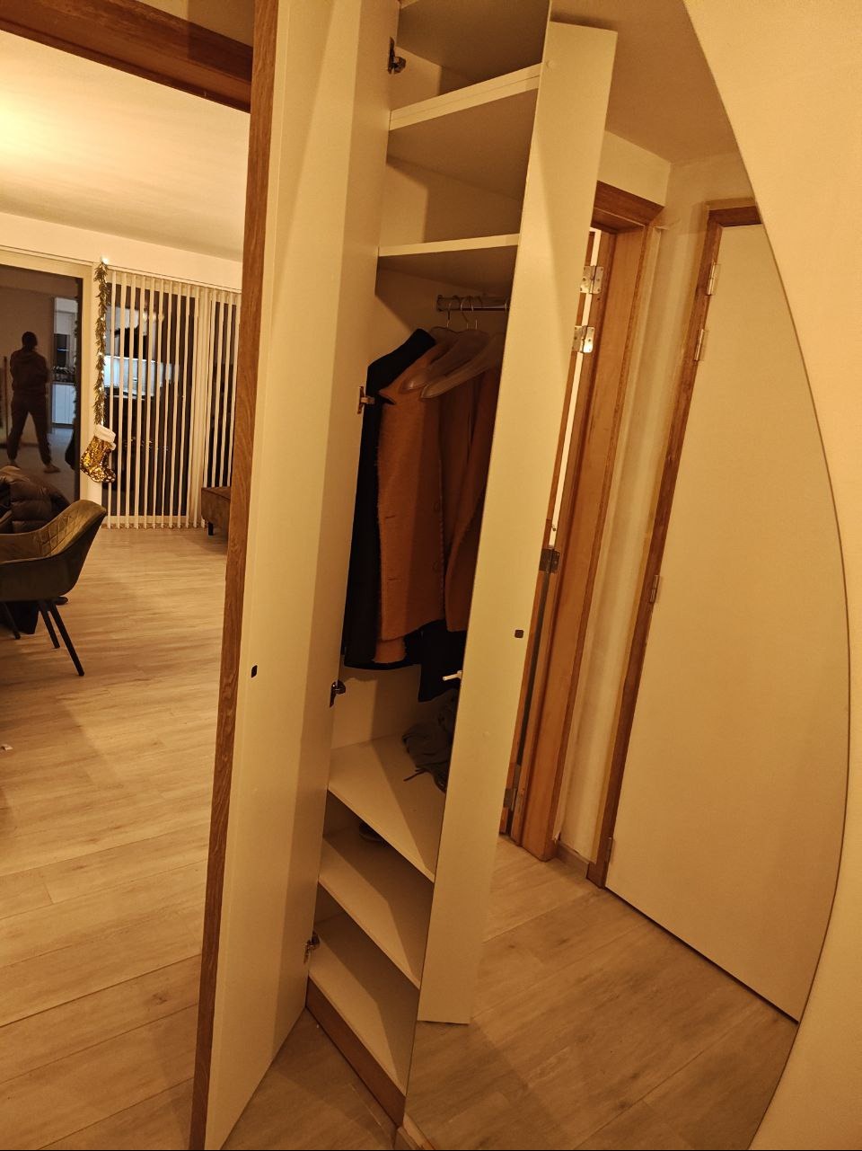 Biała szafa wnękowa z lustrem, częściowo otwarta, ukazująca półki i wiszące ubrania. Widoczne drewniane wykończenia i jasne wnętrze. Perspektywa z wnętrza mieszkania.