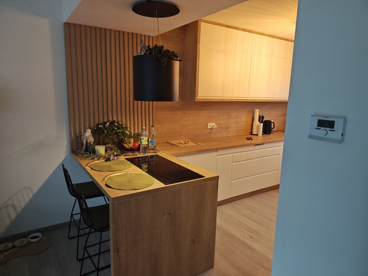 Nowoczesna kuchnia z drewnianą zabudową, wyspą i czarnym okapem cylindrycznym. Białe szafki górne, drewniany panel ścienny i dekoracje na blacie.