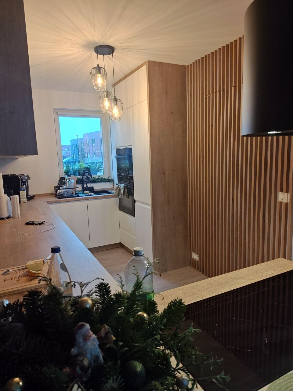 Nowoczesna kuchnia z białymi frontami i drewnianymi akcentami, oświetlona wiszącymi lampami. Ściana z drewnianych listew dodaje ciepła. Dekoracja świąteczna na pierwszym planie.