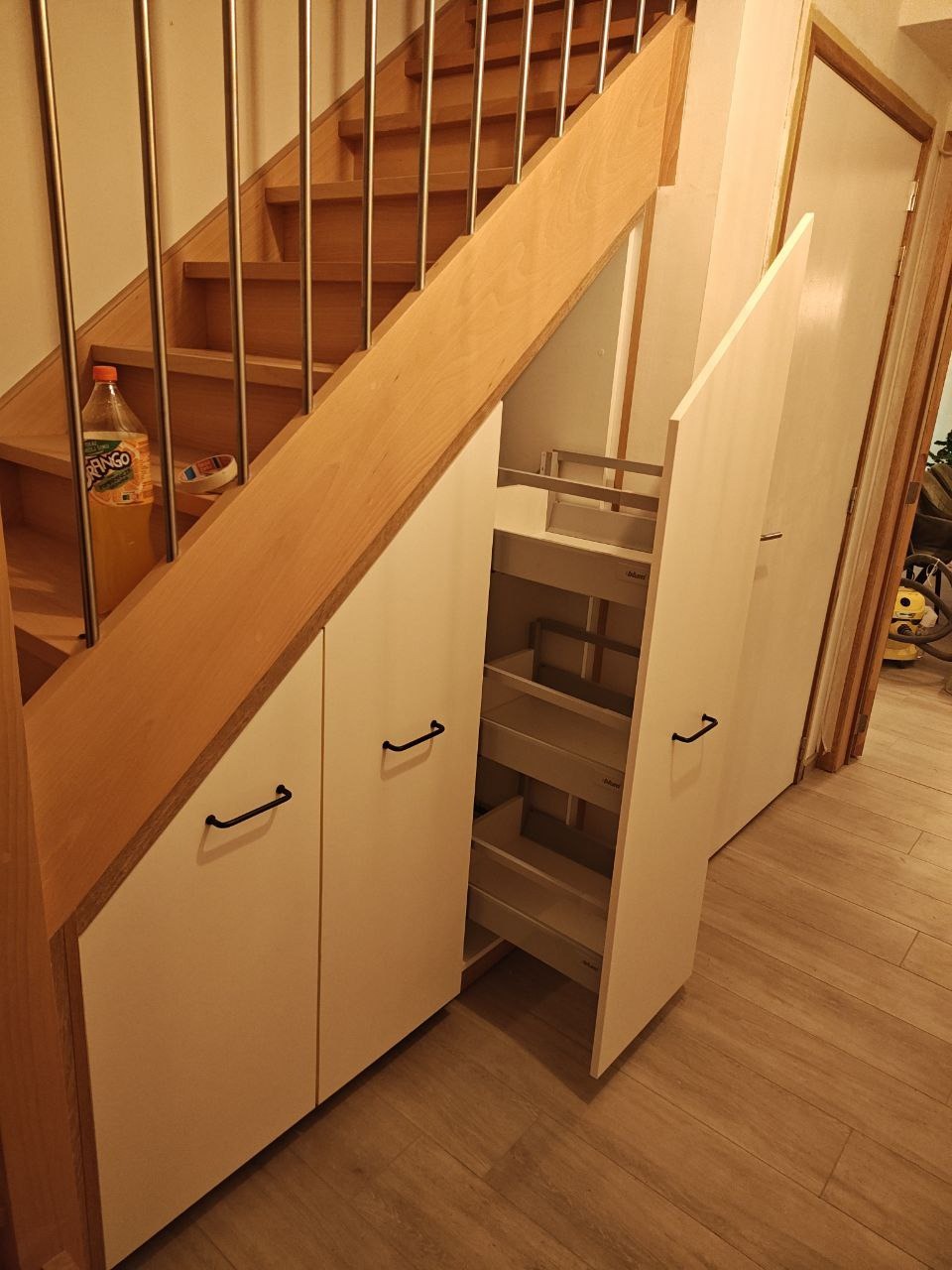Biała zabudowa pod schodami z wysuwanymi półkami, czarne uchwyty. Schody drewniane z metalową balustradą. Na schodach napój w plastikowej butelce.