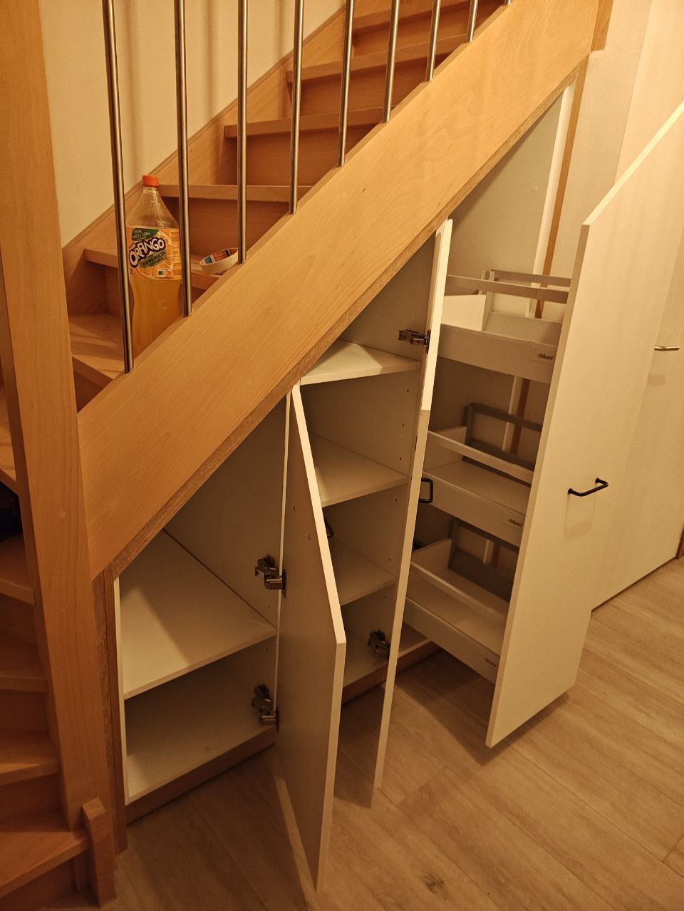 Szafka wnękowa pod schodami z białymi frontami, otwartymi drzwiczkami i wysuwanymi półkami. Na schodach stoi butelka napoju.