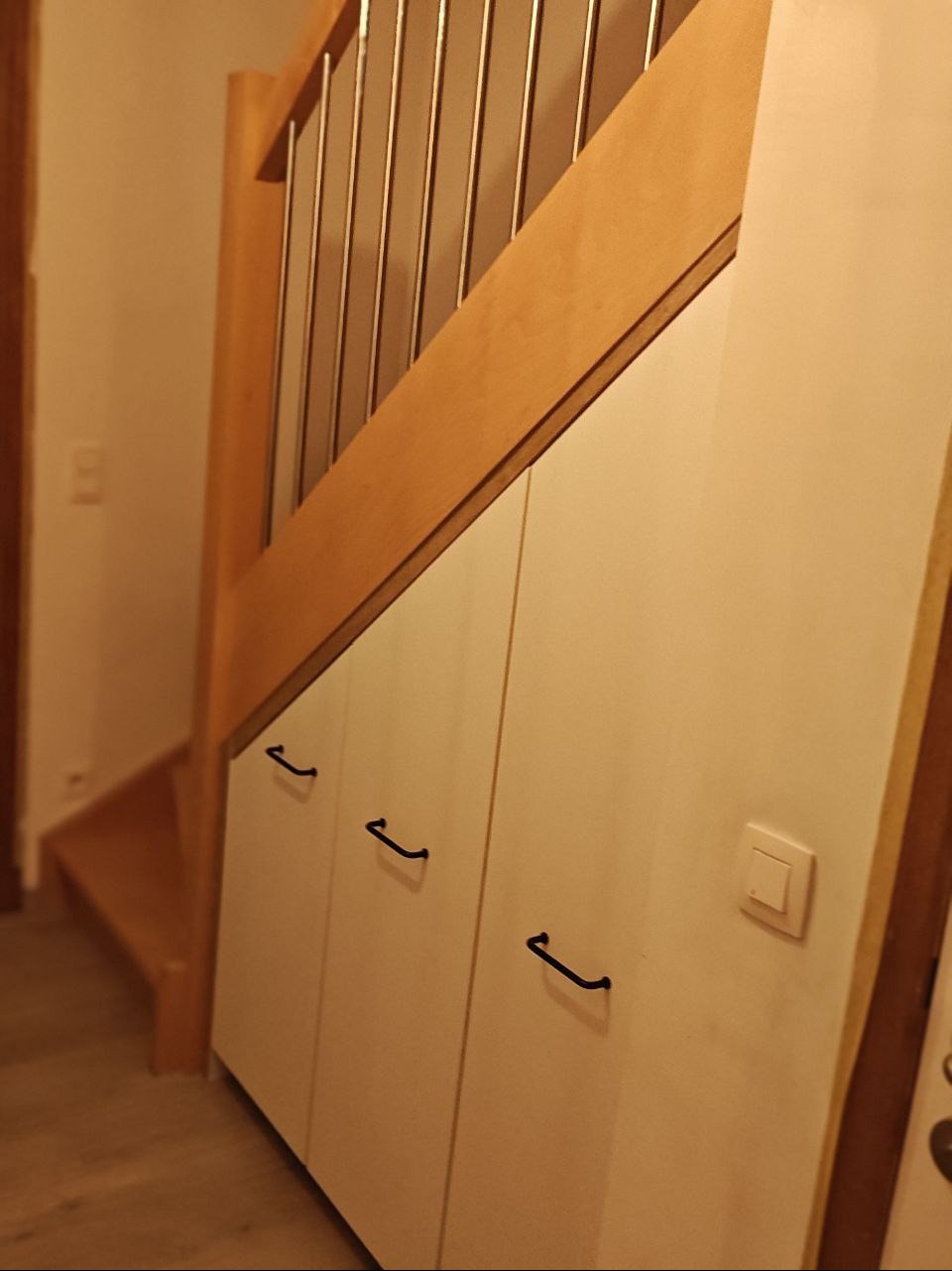 Biała szafa wnękowa pod schodami z drewnianą balustradą i czarnymi uchwytami. Widoczny fragment schodów i włącznik światła na szafie.