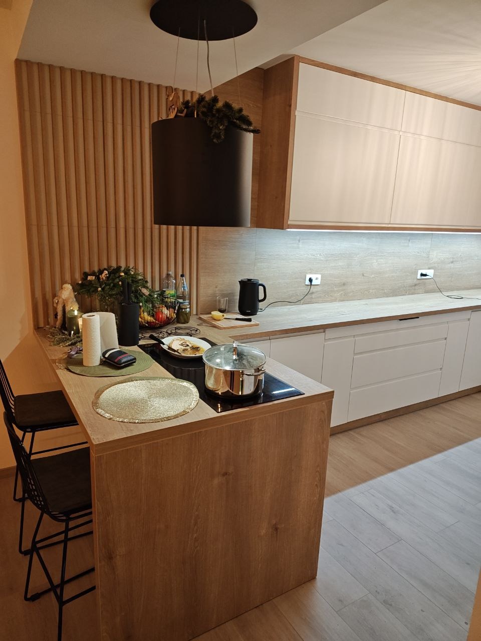 Nowoczesna kuchnia z wyspą i drewnianymi akcentami. Białe szafki wiszące, czarny okap cylindryczny i dekoracyjna ściana z drewnianych listew. Minimalistyczny design.