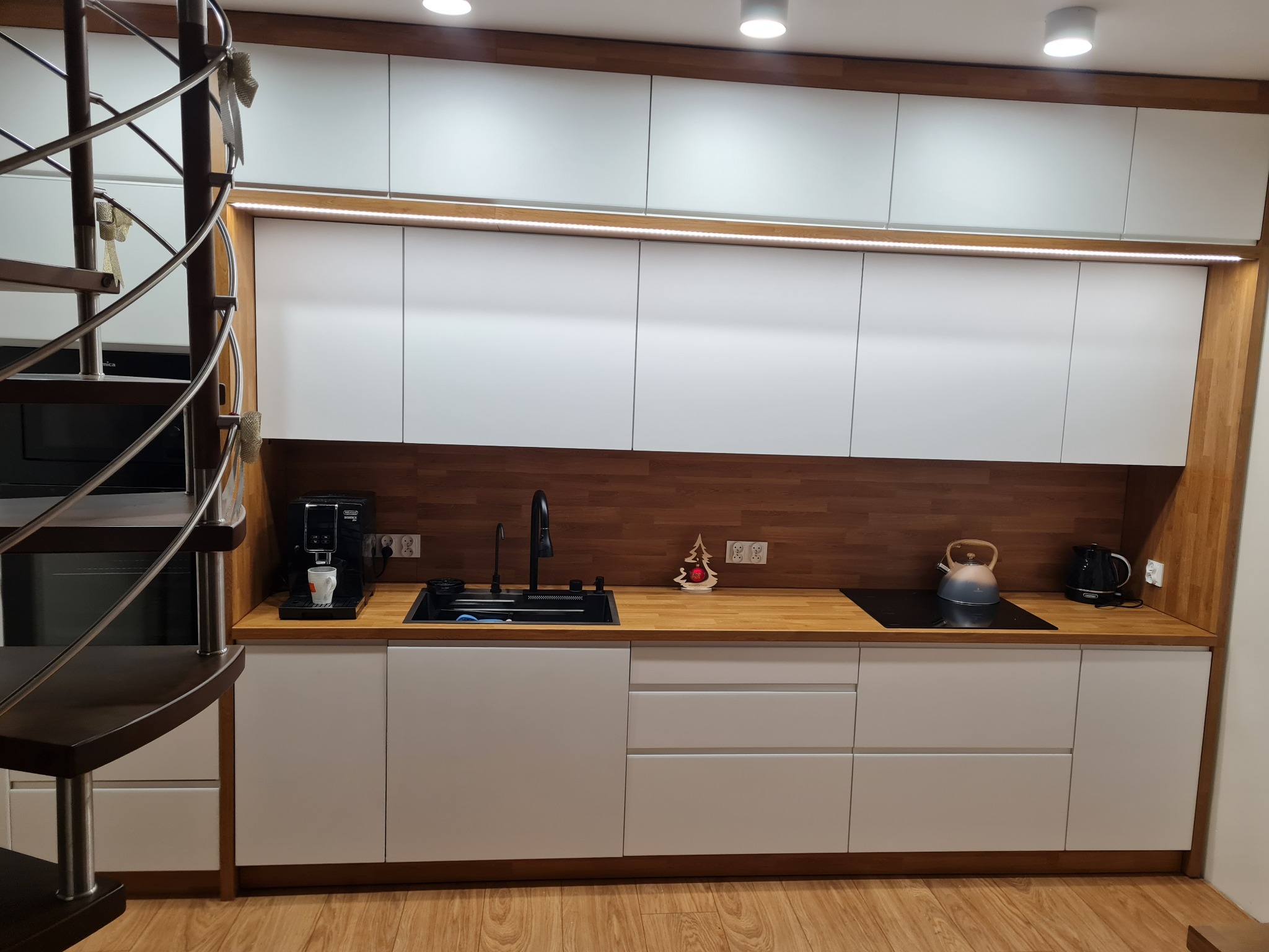 Nowoczesna kuchnia z białymi frontami i drewnianym blatem, oświetleniem LED pod szafkami wiszącymi, spiralne schody z boku. Minimalistyczny design.