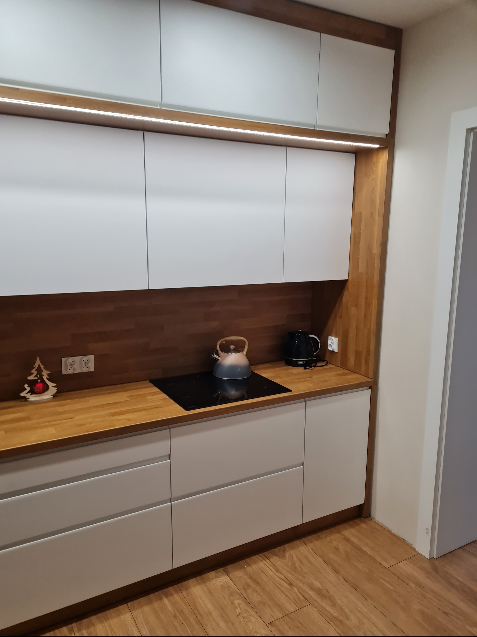 Nowoczesna kuchnia z białymi frontami i drewnianym blatem, podświetlane szafki górne, płyta indukcyjna z czajnikiem, dekoracja świąteczna.