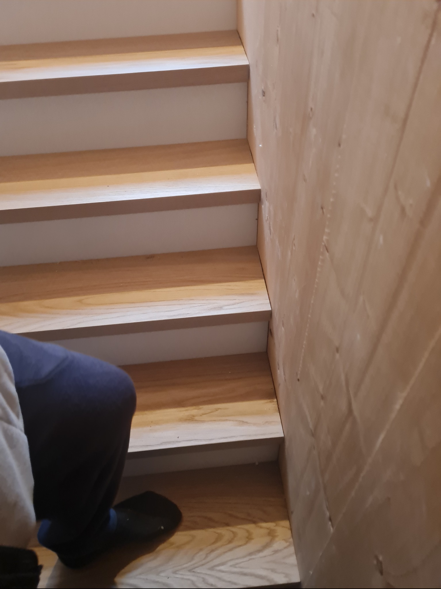 Drewniane schody z białymi podstopnicami, widok z dołu, fragment osoby w kadrze. Naturalne oświetlenie podkreśla strukturę drewna i detale wykończenia.