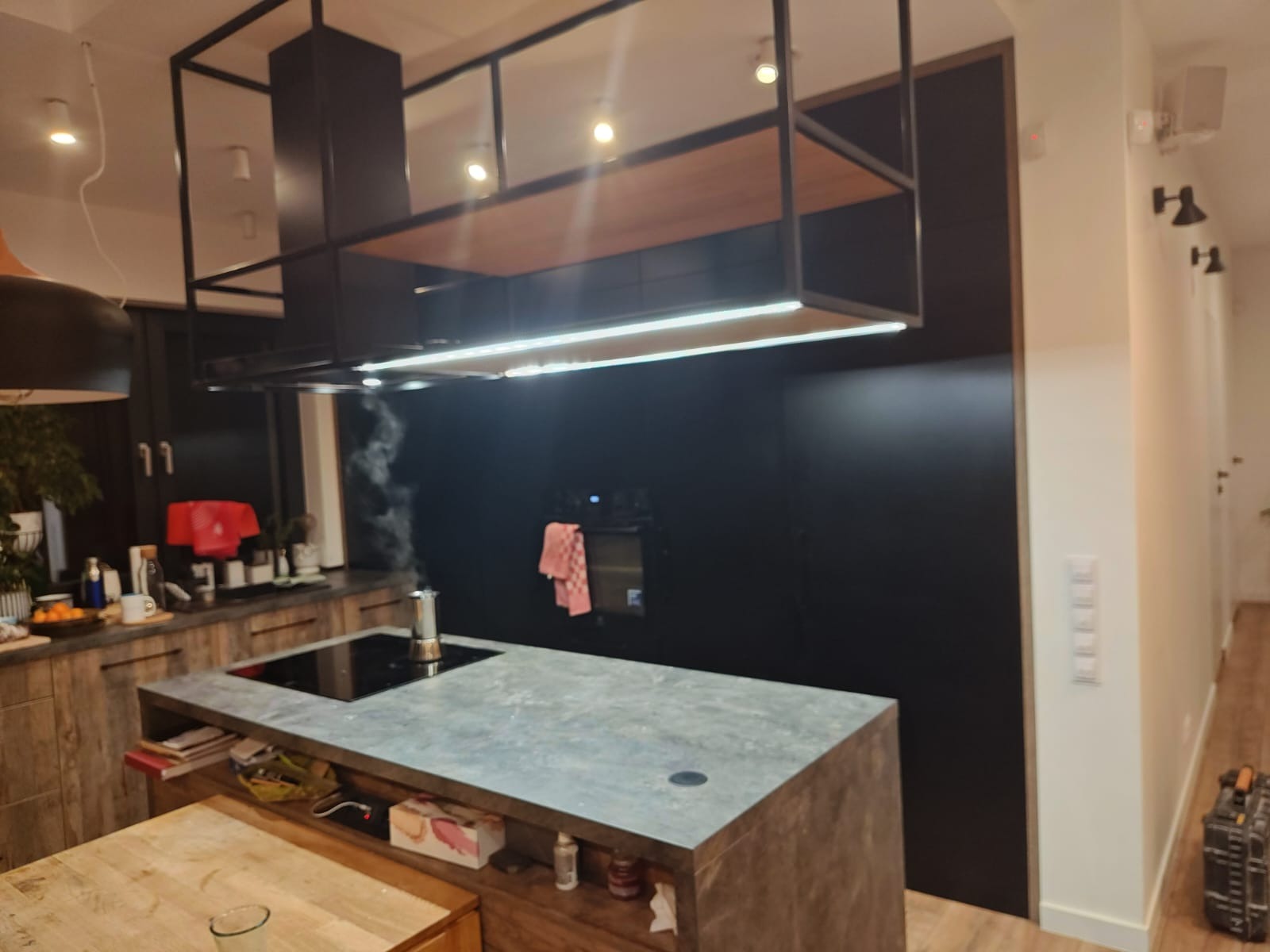 Nowoczesna kuchnia z wyspą i czarnym panelem ściennym. Nad wyspą konstrukcja z oświetleniem LED. Na blacie ekspres do kawy.