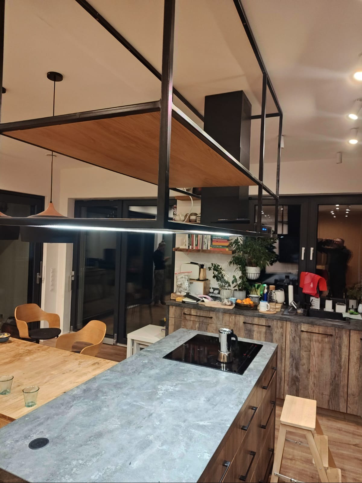 Nowoczesna kuchnia z wyspą i blatem imitującym beton. Nad wyspą konstrukcja z metalu i drewna. Widoczne drewniane szafki i półki z dekoracjami. Minimalistyczny design.