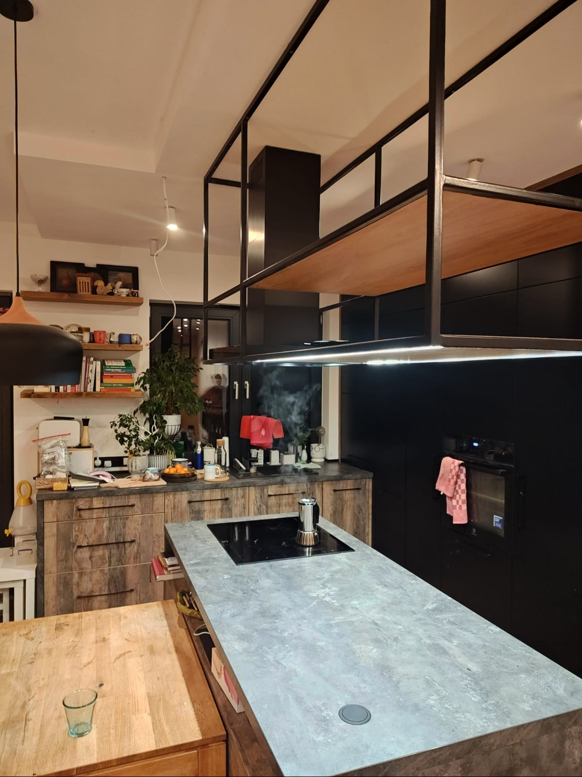 Nowoczesna kuchnia z wyspą i betonowym blatem. Na indukcji stoi zaparzacz do kawy, a nad wyspą designerski okap z drewnianą półką. Szafki w kolorze czerni i drewna.