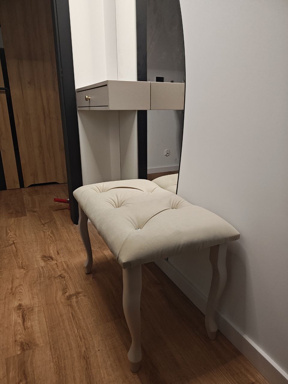 Toaletka z owalnym lustrem i tapicerowanym pufem w odcieniach beżu, na drewnianej podłodze. Widoczne detale: złoty uchwyt szuflady, dekoracyjne nóżki pufu.