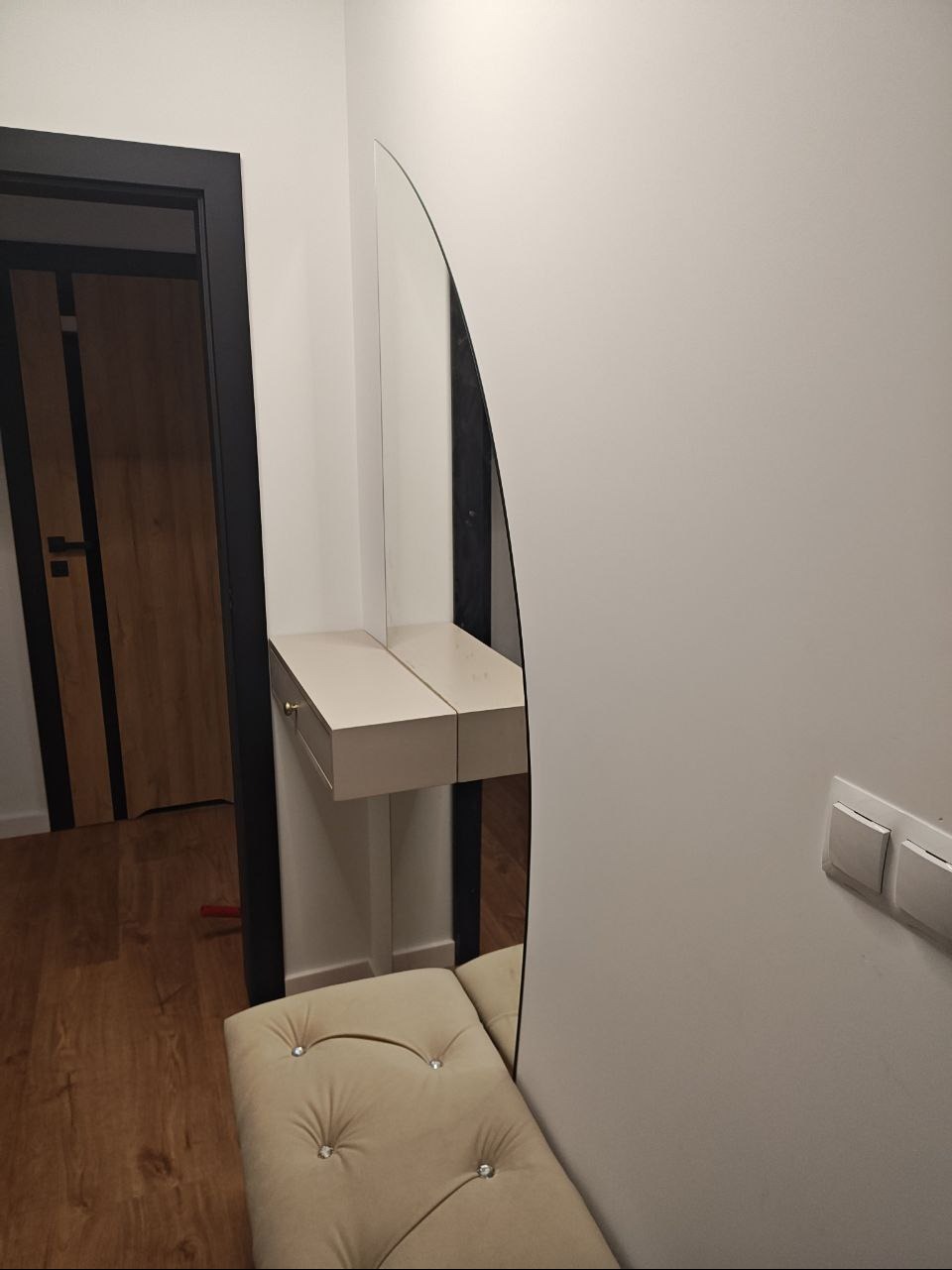 Nowoczesna toaletka z lustrem o nietypowym kształcie w przedpokoju. Beżowa pufa z guzikami i drewniana podłoga. Minimalistyczny design.