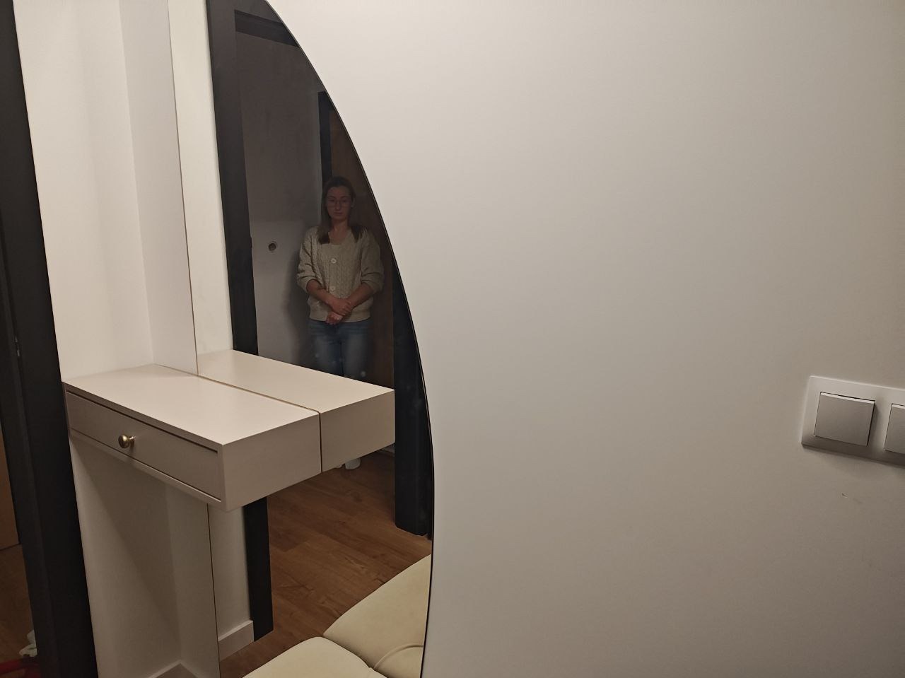 Toaletka z lustrem w kształcie łuku, odbijająca postać kobiety. Mebel w odcieniach bieli i beżu, z pojedynczą szufladą z mosiężnym uchwytem. Parkiet na podłodze.