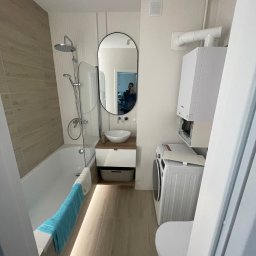 ZPRemBud Denys Shvets - Jasna łazienka z wanną, umywalką na szafce, pralką i toaletą. Na ścianach beżowe płytki imitujące drewno i białe panele. W lustrze odbicie osoby robiącej zdjęcie.