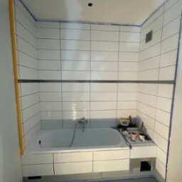 Biała wanna obudowana płytkami w łazience po częściowym remoncie. Widoczne narzędzia, poziomica, taśma malarska i fragment nieukończonej obudowy wanny.