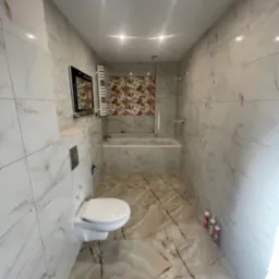 Nowoczesna łazienka po remoncie z białymi i brązowymi płytkami imitującymi marmur na ścianach i podłodze. Wisząca toaleta i grzejnik drabinkowy.